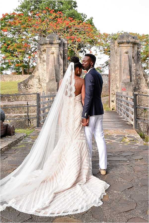 A couple shares a kiss during a bridal portrait session on a stone bridge or walkway at what appears to be a historic fort or colonial-era stone structure with large pillared gate posts. The bride wears a backless, fitted gown with geometric beaded embroidery in ivory and blush tones, paired with a very long cathedral-length veil featuring lace trim that fans out across the stone path behind her; her hair is styled in an upswept bun. The groom wears a navy blazer with white trousers and white shoes. The composition is a full-length portrait shot from behind, emphasizing the gown's dramatic train and veil against the aged stone architecture.