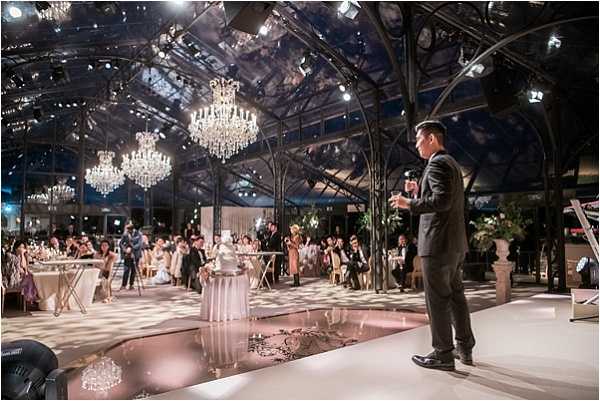 A wedding reception is underway inside a large glass-and-iron greenhouse or conservatory venue, with a vaulted transparent roof structure and ornate black metalwork arches. Multiple crystal chandeliers hang from the ceiling, providing warm ambient lighting alongside professional stage lighting rigs. In the foreground, a man in a dark suit — likely an MC or entertainer — stands on a raised stage area beside a reflective, mirrored dance floor with a pale pink tint. A large crowd of approximately 50 or more guests is seated and standing at round tables in the background, with a tiered wedding cake visible on a draped table at center. The decor palette is white and gold with greenery accents, and the overall styling is classic and formal. Wide-angle shot taken from stage level looking out toward the guests.