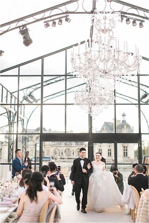 The bride and groom make their grand entrance into the reception venue, walking together down the center aisle while guests seated at long dining tables turn and photograph them on smartphones. The venue is a glass greenhouse or orangerie with black metal framing and a pitched glass roof, through which a classic French chateau — likely Vaux-le-Vicomte — is clearly visible in the background. A large crystal chandelier hangs prominently above the couple. The groom wears a black tuxedo with a bow tie, and the bride wears a strapless white ballgown with heavy lace appliqué detailing. Guests are dressed in formal attire including blush and navy. The dining tables are set with light purple or lilac linens, glassware, and what appear to be tall floral centerpieces. The overall decor style is classic and formal with a pale, neutral palette. Wide shot capturing the full room, couple, and architectural backdrop.