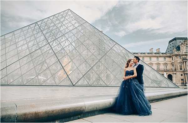 A couple poses for a pre-wedding or engagement portrait in front of the Louvre Pyramid in Paris, with the historic Louvre Palace building visible in the background. The bride wears a strapless navy blue tulle ballgown, and the groom is dressed in a matching navy blue suit, creating a coordinated color scheme. The two stand close together in an intimate embrace, leaning toward each other, with the glass-and-steel pyramid filling most of the frame behind them. This is a wide environmental portrait shot, taken at ground level, with overcast natural light.