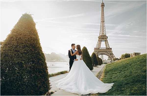 A couple poses for a wedding portrait outdoors at the Champ de Mars in Paris, with the Eiffel Tower prominently visible in the background. The bride wears a white ball gown with a long cathedral-length train, while the groom is dressed in a dark navy suit with a bow tie; they are facing each other in a close, intimate pose. The shot is a wide portrait taken from behind and to the side, emphasizing the dramatic sweep of the bride's train across the pathway, framed by manicured conical topiary hedges. The warm, golden backlit lighting suggests the image was taken during late afternoon or early evening golden hour.