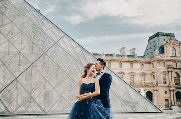 A couple poses for a pre-wedding or engagement portrait shoot in front of the Louvre Museum's glass pyramid in Paris, with the historic Louvre Palace building visible in the background. The woman wears a strapless navy blue tulle ball gown with subtle embroidered detailing, while the man wears a navy blue suit with a white dress shirt and black bow tie. He stands behind her with his arms wrapped around her waist as she smiles, and he leans in close to her face. The composition is a mid-length portrait shot with the pyramid's geometric glass-and-steel structure occupying the left portion of the frame, creating a strong diagonal line against an overcast sky.