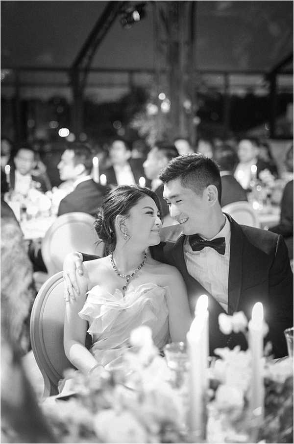 Black-and-white portrait of a bride and groom seated together at their reception dinner table, sharing a close, smiling moment. The bride wears a strapless ruffled gown with a statement necklace and an updo hairstyle, while the groom wears a dark tuxedo with a bow tie and has his arm around her. The reception appears to take place under a large tent or marquee, with candlelight and soft bokeh lighting visible in the background, where numerous seated guests fill the room. Taper candles and floral arrangements are visible in the soft-focus foreground, and the overall contrast of the image is medium with warm mid-tones suggesting candlelit ambiance.