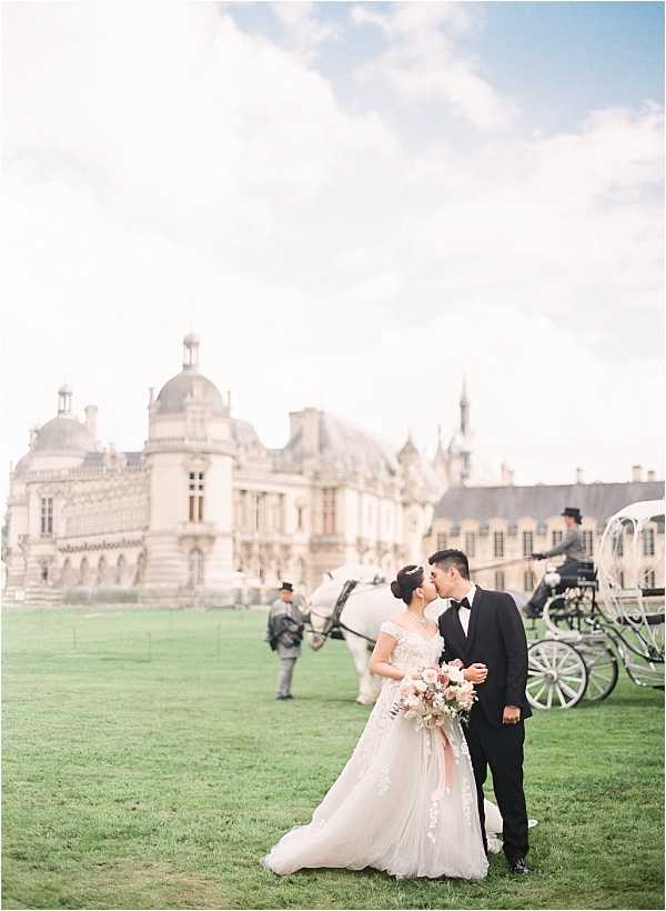A bride and groom share a kiss during an outdoor portrait session on the grounds of a large French château, identifiable by its classical French Renaissance architecture with domed rooftops and ornate stonework. The bride wears an off-the-shoulder ball gown with lace appliqué detailing and holds a bouquet of blush, dusty rose, and ivory blooms with trailing greenery, while the groom wears a black tuxedo with a black bow tie. In the background, a white horse-drawn carriage with a coachman in period dress and a second attendant on foot add to the formal, classic styling of the event. The shot is a medium-wide portrait with a softly blurred background, captured in a light, airy film-style aesthetic. Potential venue feature image.