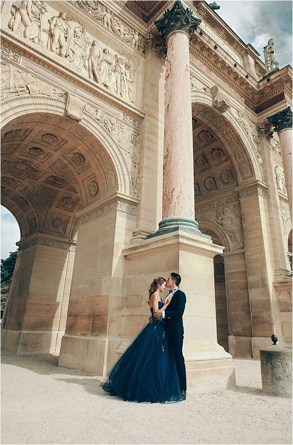 A couple shares a kiss beneath the Arc de Triomphe du Carrousel in Paris, standing at the base of a large pink marble column with ornate carved reliefs and arches visible throughout the frame. The bride wears a full-length strapless navy blue tulle ballgown, and the groom is dressed in a dark navy suit with a white dress shirt and bow tie. The shot is a wide portrait taken from ground level, emphasizing the scale of the neoclassical architecture surrounding the couple. The overall styling is formal and classic, with the bold color of the gown contrasting against the warm limestone tones of the monument.