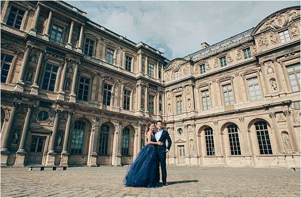 A couple poses for a portrait in the cobblestone courtyard of the Louvre museum in Paris, standing close together in front of the ornate classical French architecture. The bride wears a navy blue tulle ball gown with a fitted bodice, and the groom is dressed in a navy blue suit with a white shirt and bow tie, creating a coordinated color pairing. The wide shot frames the couple small against the grand multi-story stone facade with arched windows, decorative sculptures, and columns. Potential venue feature image.