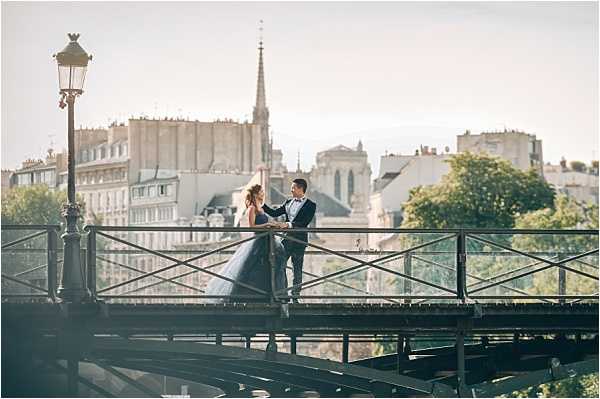 A couple poses together on a Parisian iron footbridge during what appears to be a post-wedding portrait session. The bride wears a dark navy or charcoal ballgown with a full skirt, and the groom is dressed in a dark suit with a bow tie; they face each other in a relaxed, conversational pose. The wide shot is taken from a low angle, framing the couple against a backdrop of classic Haussmann-style Parisian buildings, rooftops, and a church spire — consistent with the Île de la Cité area of Paris. A cast-iron street lamp is visible on the left side of the bridge, and the overall color palette is warm and hazy with golden late-afternoon light.