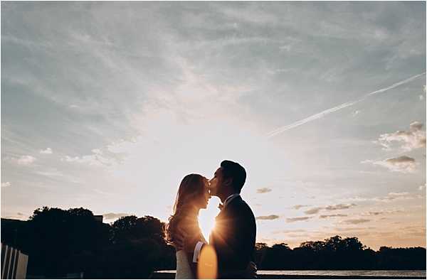 A couple shares a kiss during golden hour in a wide silhouette portrait shot, with the setting sun creating a strong backlight that renders both figures largely in shadow with warm rim lighting around their outlines. The setting appears to be an outdoor terrace or elevated platform overlooking a body of water, with a treeline visible in the background. The bride's long hair is loosely styled and the groom wears a dark suit jacket, both identifiable by their silhouettes. The composition is centered with the couple placed in the lower third of the frame against a wide expanse of open sky.