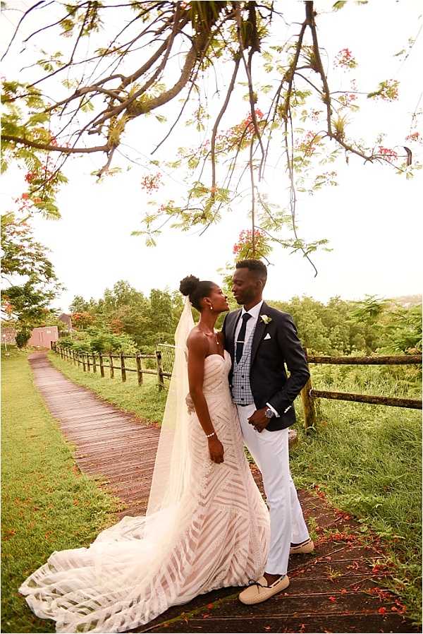 A couple portrait taken outdoors on a wooden boardwalk path lined by a rustic wooden fence, with lush green grounds visible in the background. The bride wears a strapless, fitted gown with geometric lace patterning in ivory and a long cathedral-length veil, with her hair styled in an updo; the groom kisses her cheek while wearing a dark navy blazer over a checked shirt, a dark tie with a white pocket square, white trousers, and light tan loafers. The groom has a small white boutonniere on his lapel, and fallen red flamboyant tree flowers are scattered across the boardwalk. The shot is a full-length portrait with a slightly overcast bright sky, giving soft, even lighting across the scene.