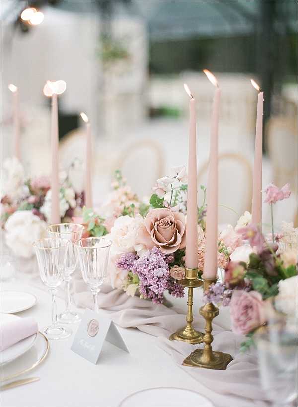 Close-up detail shot of a wedding reception tablescape featuring a soft, romantic color palette of blush, mauve, and lilac. The centerpiece includes dusty pink garden roses, lavender lilac clusters, ivory blooms, and greenery arranged in low floral clusters along a draped blush-grey chiffon table runner on a white linen tablecloth. Antique brass candlestick holders hold tall blush-pink taper candles, and crystal glassware, gold-rimmed white plates, a pale pink folded napkin, and a small grey tent place card with a wax seal are visible in the foreground. The overall styling is classic French with vintage touches, and the setting appears to be an indoor reception space with soft, diffused natural light.