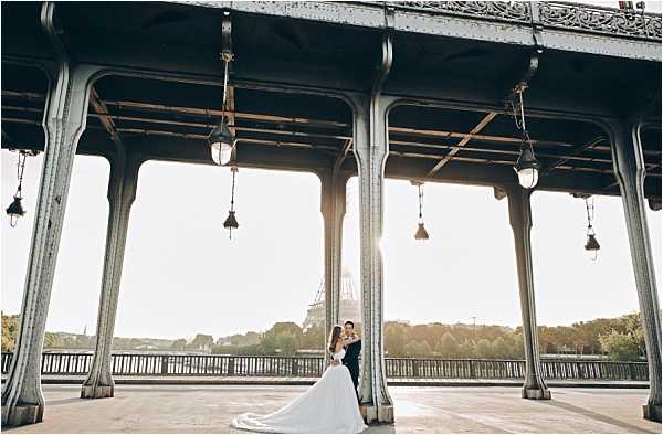 A couple poses for a portrait on the Pont de Bir-Hakeim bridge in Paris, with the Eiffel Tower visible in the background against a bright backlit sky. The bride wears a white ballgown with a full skirt and train, while the groom is dressed in a dark navy suit; the two are embracing and facing each other. The shot is taken from beneath the bridge's upper level, framing the couple between ornate cast-iron columns with hanging vintage lanterns. This is a wide environmental portrait with strong backlighting creating a sun flare effect between the architectural columns.