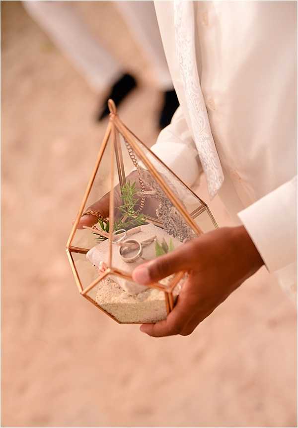 Close-up detail shot of a ring bearer holding a geometric rose gold and glass terrarium-style ring box during a wedding ceremony. The terrarium is filled with white sand, small green foliage sprigs, and holds two wedding bands alongside what appears to be a rose gold chain. The person holding it is dressed in white, and a blurred figure in black shoes is visible in the background. The styling is modern and geometric, with the rose gold metal frame forming a diamond/teardrop shape as an intentional alternative to a traditional ring pillow.