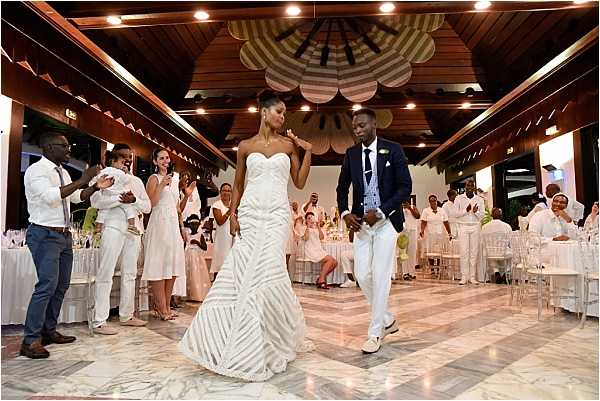 The bride and groom make their grand entrance into the reception venue, walking hand-in-hand onto a marble-tiled dance floor as approximately 30-40 guests applaud and cheer on either side. The indoor venue features a distinctive wooden ceiling with large decorative fan-shaped installations in striped cream and tan tones, recessed lighting, and a white-on-white decor palette throughout. The bride wears a strapless fitted white gown with a geometric stripe pattern and mermaid silhouette, while the groom is dressed in a navy blazer with white trousers and white shoes. Guests are dressed predominantly in white and cream attire, and the reception tables in the background are set with white linens, clear ghost chairs, and floral centerpieces with white blooms. This is a wide-angle shot capturing the full room and the energy of the entrance moment.