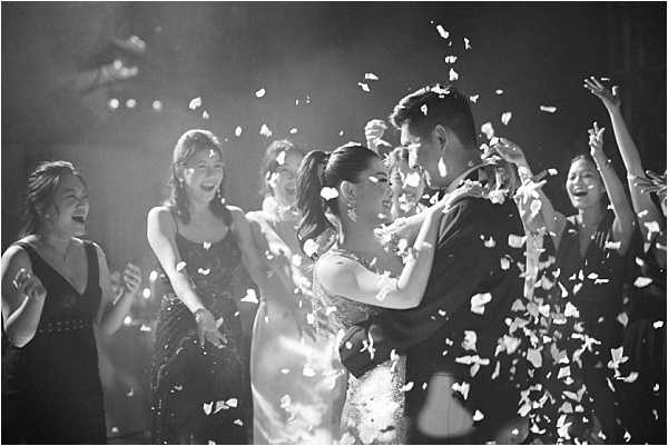A black-and-white image capturing a couple's first dance or celebratory exit as confetti or flower petals are showered around them. The bride, wearing a strapless fitted gown with her hair in an updo, embraces the groom in a dark suit as they face each other closely. Approximately five to six female guests surround them, laughing and cheering, dressed in formal gowns and cocktail dresses. The scene is lit with a strong backlight that creates bright highlights and deep contrast against a dark background, giving the image a dramatic, high-contrast look typical of indoor reception or ballroom settings.