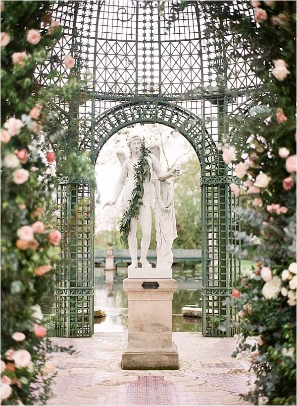 A wide shot of an outdoor garden ceremony or styled shoot setting featuring a dark green ornate iron lattice pavilion with a domed roof, framing a classical white marble statue of two figures on a stone pedestal. The statue is draped with a green garland of leaves. The pavilion columns and arches are heavily decorated with blush and pale pink roses, with lush green foliage integrated throughout the floral installations. A reflecting pool or water feature is visible through the archway in the background, and a herringbone brick pathway leads toward the statue. The overall decor palette is blush pink, ivory, and green against the dark green ironwork, creating a classic French formal garden aesthetic. Potential venue feature image.