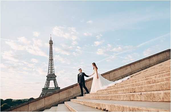 A couple poses on the wide stone steps of the Trocadéro in Paris, with the Eiffel Tower prominently visible in the background under a soft blue sky. The bride wears a flowing white gown with a long train that cascades down the steps, while the groom is dressed in a dark navy suit with a white dress shirt and black bow tie. The bride leads the groom up the steps while holding his hand, both facing each other. The shot is a wide-angle portrait taken during golden hour, capturing both the couple and the iconic Parisian landmark in a classic city elopement or post-wedding portrait session style.