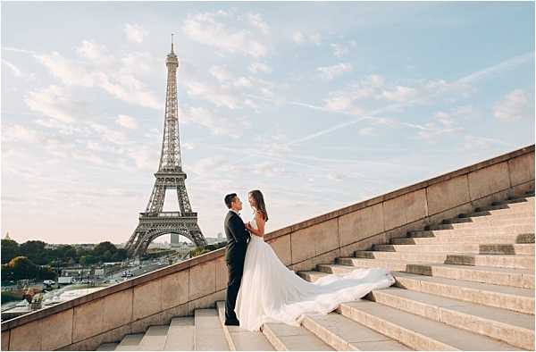 A couple poses on wide stone steps at the Trocadéro in Paris, with the Eiffel Tower prominently visible in the background. The bride wears a flowing white ballgown with a long train that cascades down the steps, while the groom is dressed in a dark navy or black suit; the two face each other closely. The shot is taken from a medium-wide angle at a low perspective, emphasizing the dramatic sweep of the gown and the monumental staircase against the open sky. The overall styling is classic and formal, with no additional bridal party or decor elements visible.