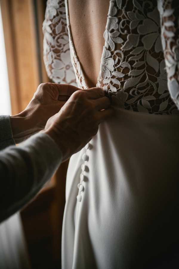 A close-up detail shot of a bride getting dressed, showing a pair of hands fastening the back of her wedding gown. The dress features a deep V-back with intricate floral lace appliqué on the bodice and a smooth ivory crepe skirt with a row of fabric-covered buttons running down the center back. The shot is tightly cropped, focusing on the hands and the back of the dress, with warm, soft natural light coming from a window in the background. The styling is classic and refined, with the contrast between the textured lace bodice and the clean lines of the skirt being a clear intentional design feature.