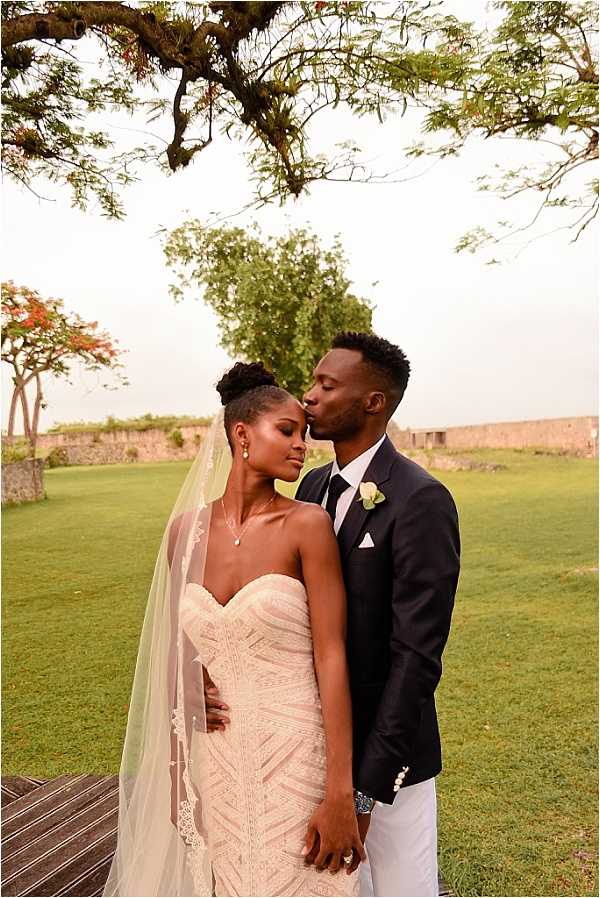A couples portrait taken outdoors on a manicured lawn, with the groom gently kissing the bride's temple as she closes her eyes. The bride wears a fitted, strapless ivory lace gown with intricate geometric beading and a long cathedral-length veil with lace trim, paired with a delicate pendant necklace and drop earrings; her hair is styled in an updo. The groom wears a navy blazer with white trousers, a white dress shirt, and a cream boutonniere. The setting appears to be the grounds of a historic fortified structure, with low stone walls visible in the background. The shot is a mid-length portrait with soft, warm late-afternoon light.