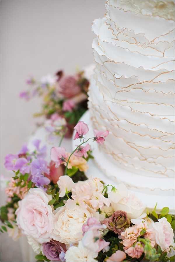 Close-up detail shot of a multi-tiered white wedding cake decorated with layered fondant ruffles trimmed with fine gold-painted edges. The base of the cake is surrounded by a lush arrangement of fresh flowers in blush pink, soft lavender, mauve, and cream tones, including garden roses, peonies, sweet peas, and ranunculus with green foliage. A second floral cluster is softly out of focus in the background. The overall palette is romantic and soft, consistent with a classic French wedding aesthetic.