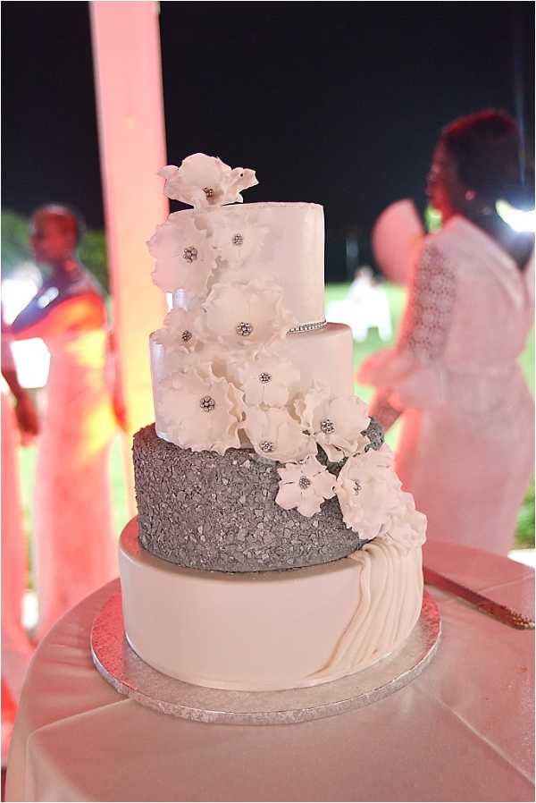 Close-up detail shot of a four-tier wedding cake displayed on a silver cake board over a white satin-draped table, photographed at an outdoor evening reception. The bottom tier features ivory fondant with vertical pleating, the second tier is covered in silver chunky glitter or crushed sequin texture, the third tier is smooth ivory fondant with a rhinestone band, and the top tier is smooth ivory. A cascading arrangement of white sugar flowers with rhinestone centers runs from the top to the bottom of the cake. In the blurred background, guests in formal attire are visible under pink ambient uplighting against a dark night sky.