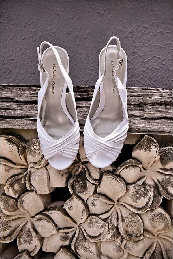 A detail close-up shot of a pair of white satin bridal heels with crossed straps, a peep toe, and slingback ankle straps, resting on a weathered wooden ledge above a decorative stone or concrete panel featuring a repeating floral relief pattern. The insoles are branded with what appears to read 'Colorful Creations.' The shoes are white against a textured grey wall background, with the stone floral panel in muted beige and grey tones beneath them. The styling is classic bridal.