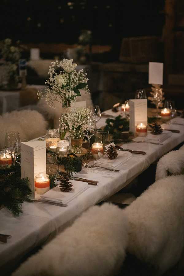 A close-up detail shot of a wedding reception tablescape set for an indoor evening dinner, styled with a rustic winter theme. The long table is dressed with a white linen runner and decorated with mason jar arrangements of baby's breath and white blooms, scattered pinecones, fresh evergreen garland, and multiple lit votive and pillar candles in amber and white holders that cast warm, low light across the setting. Place settings include silver cutlery, folded white napkins, wine glasses, and small white rectangular menu or place cards. The bench seating on either side is covered with fluffy ivory sheepskin or faux-fur cushions, reinforcing the cozy, alpine-chalet aesthetic.