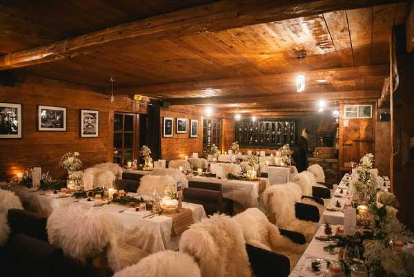 Indoor wedding reception setup inside a rustic alpine chalet or mountain lodge with exposed dark wood beam ceilings and wood-paneled walls. The room is arranged with multiple long rectangular tables covered in white linens, with white sheepskin or faux fur throws draped over each chair. Table centerpieces feature baby's breath, small white floral arrangements, and clusters of candles and tea lights in glass vessels creating warm ambient lighting throughout the room. The decor palette is white, cream, and gold against the dark wood surroundings, with greenery runners along the tables and black-and-white framed photographs hanging on the walls. Wide shot taken from the entrance end of the room, showing approximately 10–12 tables with seating for an estimated 60–80 guests, with no people present.