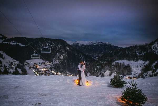 A couple shares a close embrace on a snowy mountain slope at dusk, with a ski resort village visible in the valley below and a chairlift cable running overhead. The bride wears a white long-sleeve fitted gown and the groom wears dark trousers with a waistcoat. Two warm-glowing lanterns are placed in the snow at their feet, providing the primary light source in the near-dark conditions. The wide-shot composition captures a winter alpine wedding portrait setting with deep blue twilight tones across the sky and mountain range.