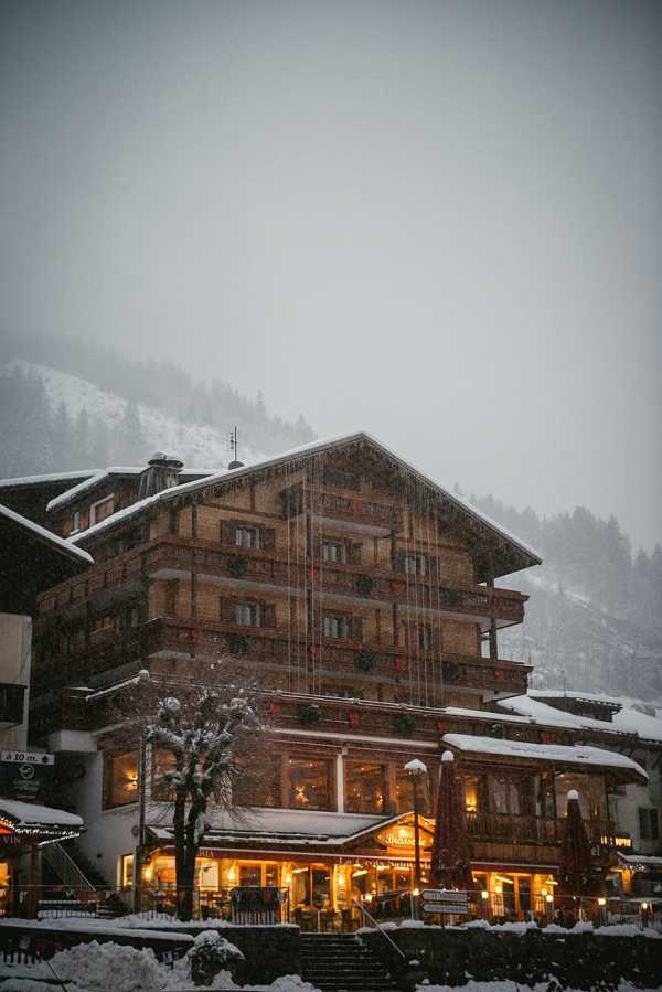 Wide exterior shot of a large Alpine chalet-style hotel building photographed during heavy snowfall at dusk or early evening. The multi-story timber and stone building features warm amber and golden interior lighting glowing from its ground-floor restaurant and bar area, with a sign partially readable as 'La Croix' visible on the facade. The building is decorated with greenery and red accents on the balconies, consistent with winter holiday styling. No people, couples, or wedding activity are visible in the frame. Potential venue feature image.