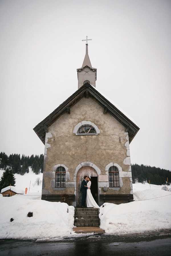 A couple shares a kiss on the front steps of a small Alpine chapel in a winter mountain setting, with deep snow surrounding the building on all sides. The bride wears a long-sleeved white gown with a flowing train, and the groom is dressed in a dark suit. The chapel features rendered stone walls, arched windows with white stone surrounds, a wooden steeple, and a cross at the top. This is a wide portrait shot taken straight-on, emphasizing the chapel's symmetrical facade with the couple centered in the doorway. Potential venue feature image.