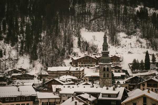 Wide-angle landscape shot of a snow-covered Alpine village in winter, featuring a prominent stone church tower with a pointed steeple at the center, surrounded by traditional chalet-style buildings with snow-laden rooftops. A forested mountainside rises steeply behind the village, with bare deciduous trees and snow between them. The image has a muted, cool tone with overcast lighting. No people, couples, or wedding-specific elements are visible in the frame. Potential venue feature image.
