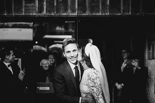 This black-and-white portrait captures the couple just after what appears to be a ceremony exit, with the bride leaning in to kiss or whisper to the groom as he smiles broadly at the camera. The groom wears a dark suit with a tie, while the bride is dressed in a long-sleeved lace gown and a long veil. Several guests are visible in the background watching and photographing the moment, set against the exterior of what appears to be a stone or rendered building. The image has strong contrast with deep shadows and bright midtones, shot at close-to-medium portrait distance.