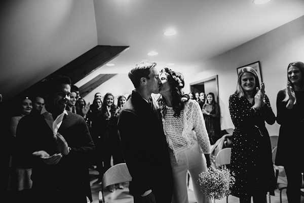 A black-and-white image capturing the couple's first kiss during an indoor civil ceremony, set in a small room with a sloped ceiling and recessed lighting. The bride wears a long-sleeved lace or crochet-detail dress and has a floral crown in her hair, holding what appears to be a small bouquet of baby's breath; the groom is in a dark suit. Approximately 15–20 guests line both sides of the aisle, many applauding and smiling, with strong contrast between the bright central figures and the darker surrounding crowd. The composition is a mid-range portrait shot taken from the aisle level, with the couple centered and guests framing them on either side.