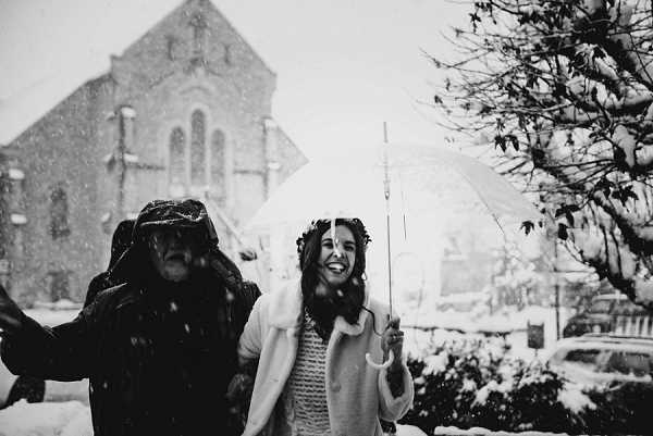A black-and-white portrait of a bride and one other person, likely a guest or companion, standing outside a stone church in heavy snowfall. The bride is laughing and holding a clear bubble umbrella, wearing a light-colored coat over her wedding outfit and a floral or leaf crown in her dark hair. The companion beside her is dressed in a dark hooded jacket and also appears to be reacting to the snow. The image has high contrast with bright whites from the falling snow against the mid-toned stone church façade visible in the background. Medium-distance two-person shot with a candid, spontaneous feel.
