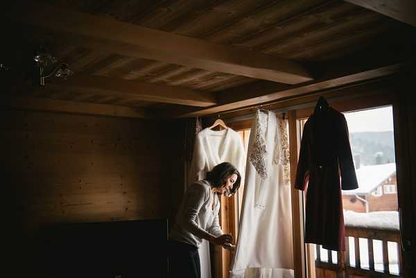 A bride is getting ready in a dimly lit wooden chalet room, standing near a window with a snowy mountain scene visible outside. Two garments are hanging in the window frame: a simple long-sleeve white cover-up or second dress, and a heavily embellished ivory wedding gown with lace or beaded detailing, alongside a dark robe. The bride, dressed casually in a grey top, appears to be fastening or adjusting something at her waist. The room features dark wood-paneled walls and ceiling, consistent with an Alpine chalet setting, creating a moody, low-light atmosphere. Wide portrait shot capturing the getting-ready moment.