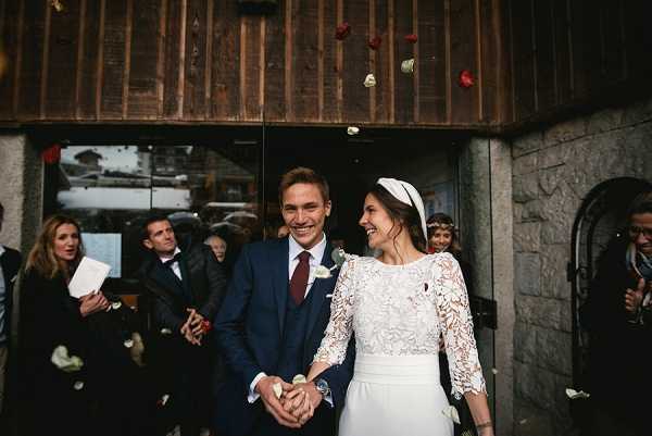 The bride and groom exit a ceremony venue through a dark wooden doorway as guests toss red and white flower petals over them. The groom wears a navy three-piece suit with a burgundy tie and a white boutonniere, while the bride wears a long-sleeved lace top with a simple white skirt and a white structured headband. Approximately eight to ten guests are visible in the background, some holding order-of-service booklets. The shot is a mid-range portrait capturing the couple laughing and holding hands, with a rustic stone and dark timber building facade suggesting an alpine or mountain village setting.