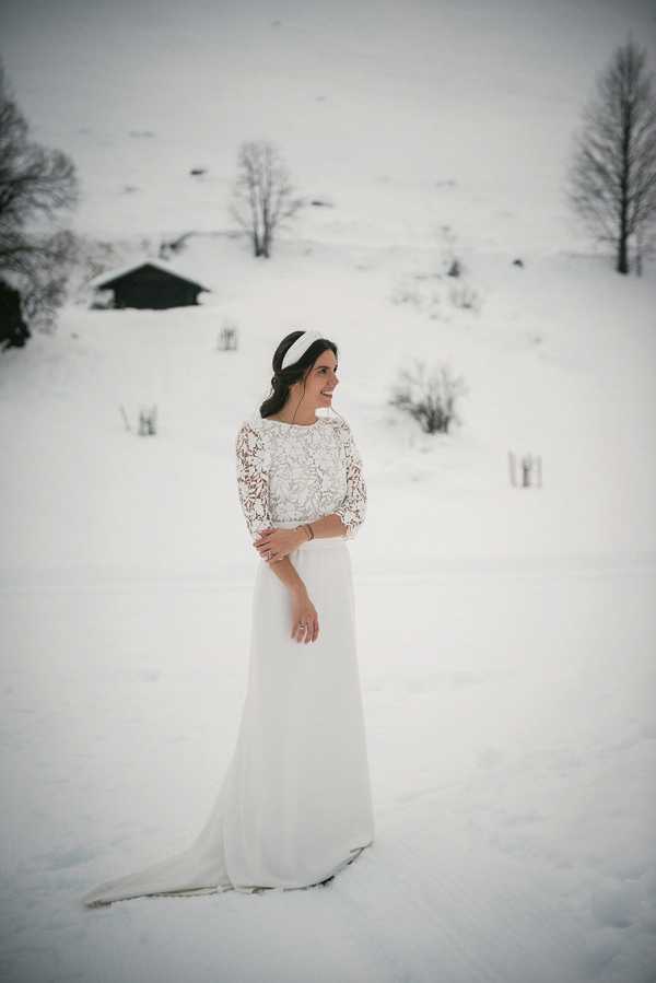 A bridal portrait taken outdoors in a snow-covered winter landscape, with a dark chalet structure and bare trees visible in the background. The bride stands alone, smiling and looking off to one side, wearing a two-piece ivory gown with a lace three-quarter-sleeve top and a plain flowing skirt with a short train. She accessorizes with a wide white fabric headband and a bracelet. The image has a muted, slightly desaturated tone consistent with winter light. Medium full-length portrait composition.