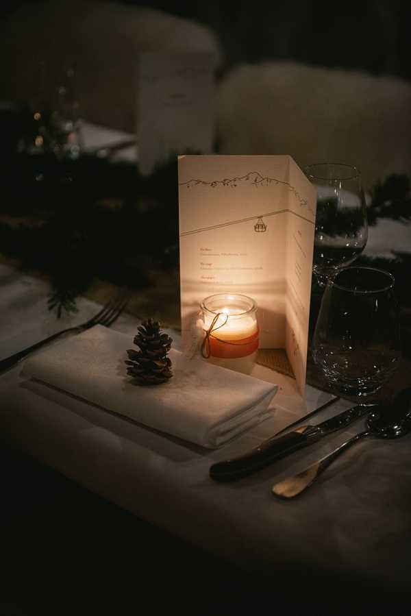 Close-up detail shot of a single place setting at a winter-themed wedding reception, photographed in low candlelight. The setting includes a white folded napkin topped with a small pine cone, a lit amber glass votive candle that illuminates a tri-fold menu card illustrated with a mountain skyline and cable car motif, and two clear wine glasses alongside dark-handled cutlery. Greenery consistent with pine or fir branches is visible in the background as part of the table centerpiece. The overall decor palette is dark and moody with warm amber candlelight, and the styling theme is alpine or mountain rustic, likely for a winter wedding.