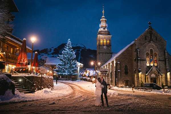 A couple shares an embrace in the middle of a snow-covered village square during a winter evening, shot as a wide environmental portrait. The bride wears a white gown and the groom is in dark formal attire. The square features a lit Christmas tree decorated with white lights at its center, flanked by a stone church with an illuminated bell tower on the right and a chalet-style building with red patio umbrellas on the left. Street lamps cast a warm amber glow across the snow, and the deep blue of the night sky creates strong contrast with the lit surroundings. The overall styling is a winter alpine wedding with a festive holiday atmosphere.