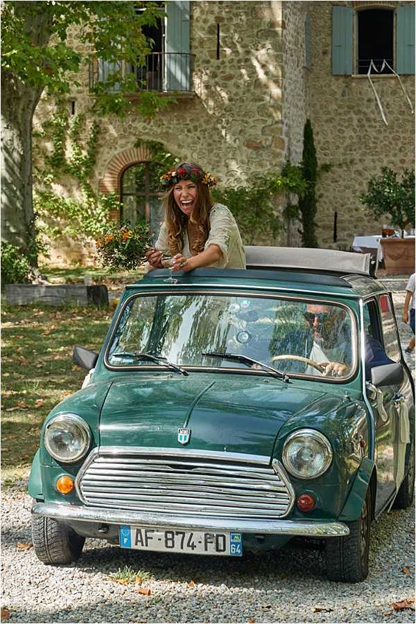 A bride leans out of the open sunroof of a vintage teal-green classic Mini Cooper, laughing and holding a small bouquet of orange and yellow wildflowers. She wears a floral crown with deep red and orange blooms, a relaxed bohemian-style cream top, and her long wavy hair falls loosely around her shoulders. The groom sits in the driver's seat wearing sunglasses, partially visible through the windshield. The setting is the gravel courtyard of a stone-facade French country house or mas, with teal-painted shutters visible on the building facade behind them. The overall styling is relaxed and boho. Medium-distance portrait shot taken from ground level, front-facing the vehicle.