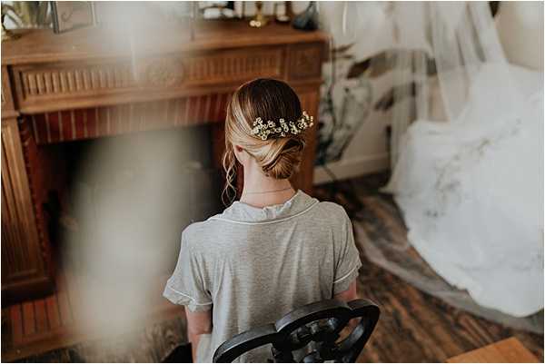 A getting-ready shot taken indoors, showing the back of a bride seated on a dark wooden chair in front of a warm-toned wooden fireplace mantel. She is wearing a casual grey t-shirt and her hair is styled in a low chignon adorned with small white and yellow floral hair accessories, likely baby's breath or similar delicate blooms. A white wedding dress on a hanger is partially visible at the right edge of the frame. The composition is a close-up portrait from behind, with a slightly soft, hazy quality around the edges, giving the image a candid, intimate feel.