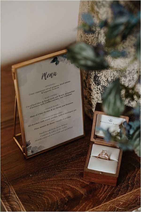 A detail flat lay shot on a wooden surface featuring a gold-framed menu card with navy blue botanical watercolor accents, displaying a multi-course French wedding menu with sections for appetizer, main course, cheese plate, and desserts. Beside it sits an open walnut wood ring box branded 'Brilliant Earth' containing a solitaire diamond engagement ring with a gold band set in white velvet lining. In the upper right corner, out-of-focus foliage with dusty blue and green tones provides a soft natural backdrop. The overall styling palette combines warm wood tones, gold accents, and muted navy botanicals in a modern rustic style.