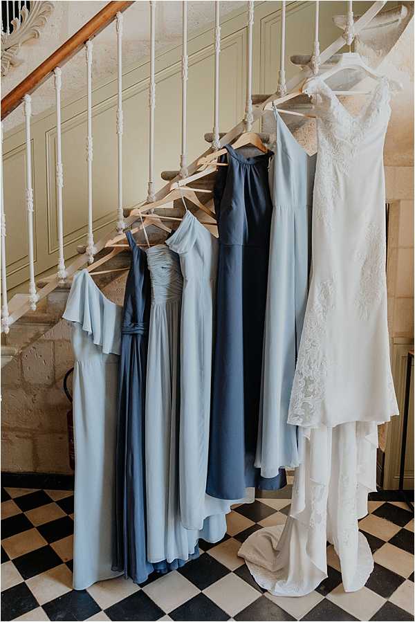 A getting-ready detail shot showing five dresses hung from the spindles of an ornate interior staircase. Four bridesmaid dresses in varying shades of blue — including dusty blue, steel blue, and navy — are displayed in full-length silhouettes with different neckline styles including flutter sleeves, halter, and ruched bodice. A white lace bridal gown with a V-neckline, lace appliqué detailing, and a trailing skirt hangs separately to the right. The dresses are hung on wooden hangers against a classic interior with panelled sage-green walls and a black-and-white chequerboard floor, suggesting a French chateau setting. The composition is a wide vertical shot capturing all garments in full length.