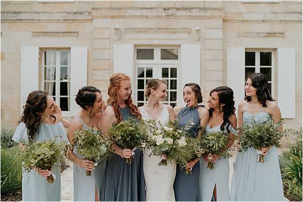 A bridal party portrait taken outdoors in front of a classic French chateau with limestone facade and white shuttered windows. The bride, dressed in a white lace gown, stands at the center surrounded by six bridesmaids wearing mismatched dusty blue and slate blue dresses in varying styles including halter neck, off-shoulder, and flutter sleeve silhouettes. All seven women hold loose, garden-style bouquets of lush greenery with white blooms, including what appears to be white ranunculus and eucalyptus. The group is laughing and interacting naturally with one another. The overall styling palette is cohesive in soft blue-grey tones with a relaxed, organic floral aesthetic. Medium-distance portrait shot with the chateau architecture as the backdrop.