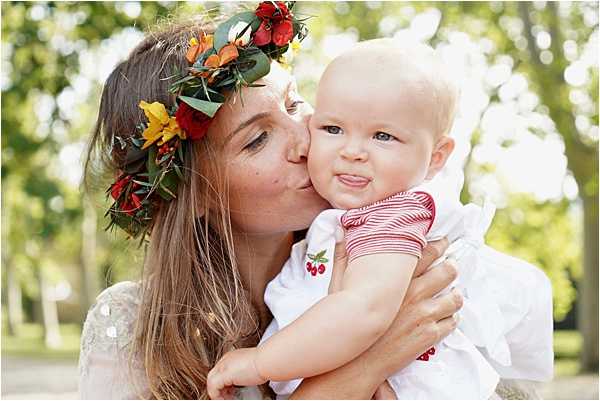 A close-up portrait of a woman, likely the bride, kissing the cheek of a baby she is holding. The woman wears a large floral crown made of red, orange, and yellow blooms with green foliage, and a white dress with subtle texture visible at the shoulder. The baby is dressed in a white outfit with red and white striped sleeves and a small cherry embroidery detail. The shot is taken outdoors in what appears to be a tree-lined setting, with soft, blurred greenery in the background. The styling has a boho feel, with the bold floral crown as the key decorative element.