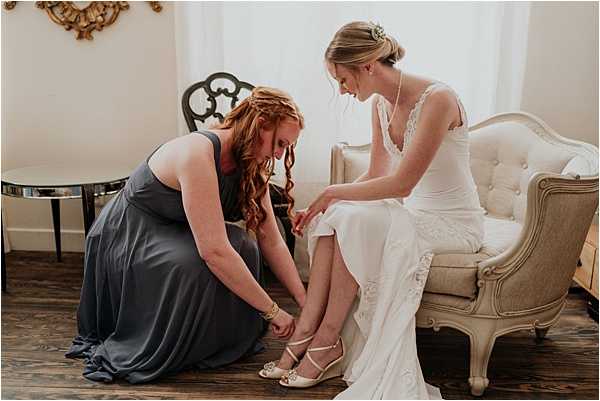 A getting-ready scene shot indoors, showing a bridesmaid helping the bride put on her shoes. The bride is seated on a cream tufted French-style settee wearing a white lace spaghetti-strap gown with a delicate pearl necklace and a floral hair accessory, while the bridesmaid, dressed in a slate blue floor-length gown, kneels on the dark hardwood floor to fasten the bride's ivory strappy wedge heels. The room features a gold ornate mirror frame on the wall and a mirrored side table, suggesting a classic, vintage-inspired interior decor style. The composition is a medium portrait shot with warm natural light coming from a window in the background.