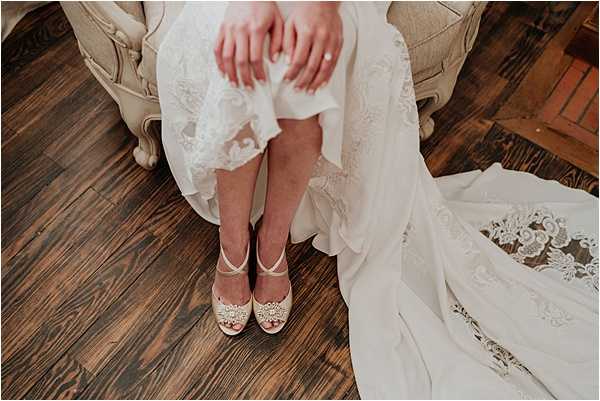 A close-up detail shot of a bride seated indoors on a cream upholstered chair, showing her lower body and feet. She is wearing a white lace wedding dress with a flowing skirt and lace appliqué train that pools on the dark hardwood floor. Her shoes are ivory open-toe heels with cross-strap ankle straps and an ornate crystal and pearl brooch detail at the toe. Her hands rest on her lap, folded over the lace skirt. The composition is a top-down portrait-style detail shot emphasizing the shoes, dress texture, and warm-toned wood flooring.