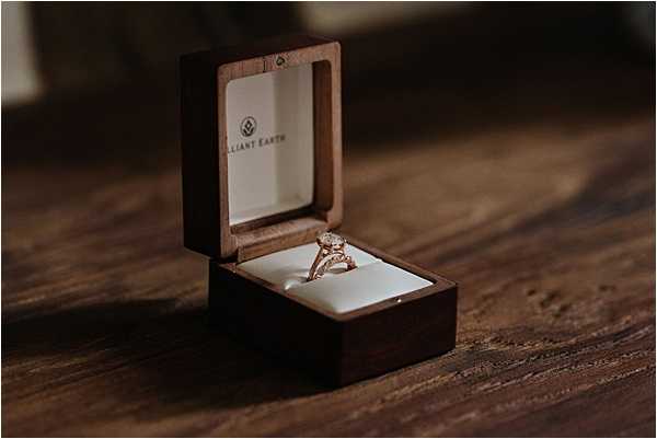 Close-up detail shot of an engagement ring displayed in an open dark walnut wood ring box branded with 'Brilliant Earth,' placed on a wooden surface. The ring features a rose gold band with a round solitaire diamond and decorative side detailing. The box interior is lined with cream-white cushioning, and the image is lit with warm, dim ambient light creating deep shadows around the subject.