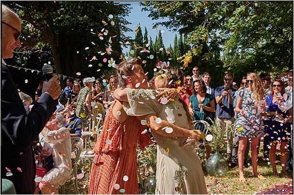A couple shares an embrace and kiss at the end of an outdoor wedding ceremony while guests throw large multicolored paper confetti discs in pastel pinks, greens, yellows, and white. The bride wears a white embroidered or lace-detailed gown, and her partner is dressed in a rust-orange or terracotta midi dress. Approximately 30 guests line both sides of the aisle, many smiling and photographing the moment, dressed in colorful summer attire. The setting is a sunny outdoor ceremony space with trees providing a backdrop, and a celebrant holding a microphone is visible on the left. The composition is a mid-range action shot capturing the confetti in motion.